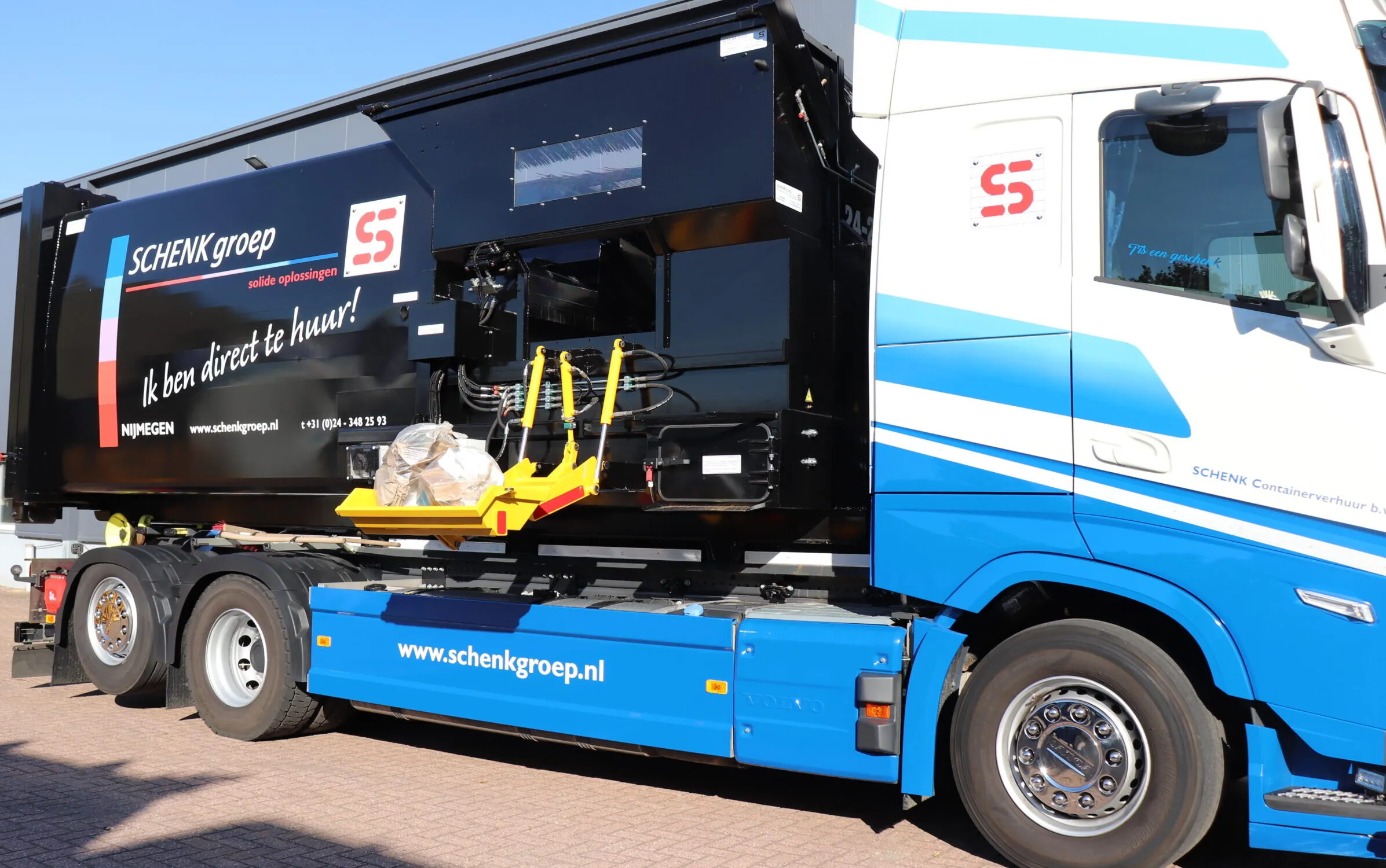 Gele en rode mechanische arm op een IPC-truck, in actie bij het beladen van afvalcontainers, met bedieningspaneel op de zijkant