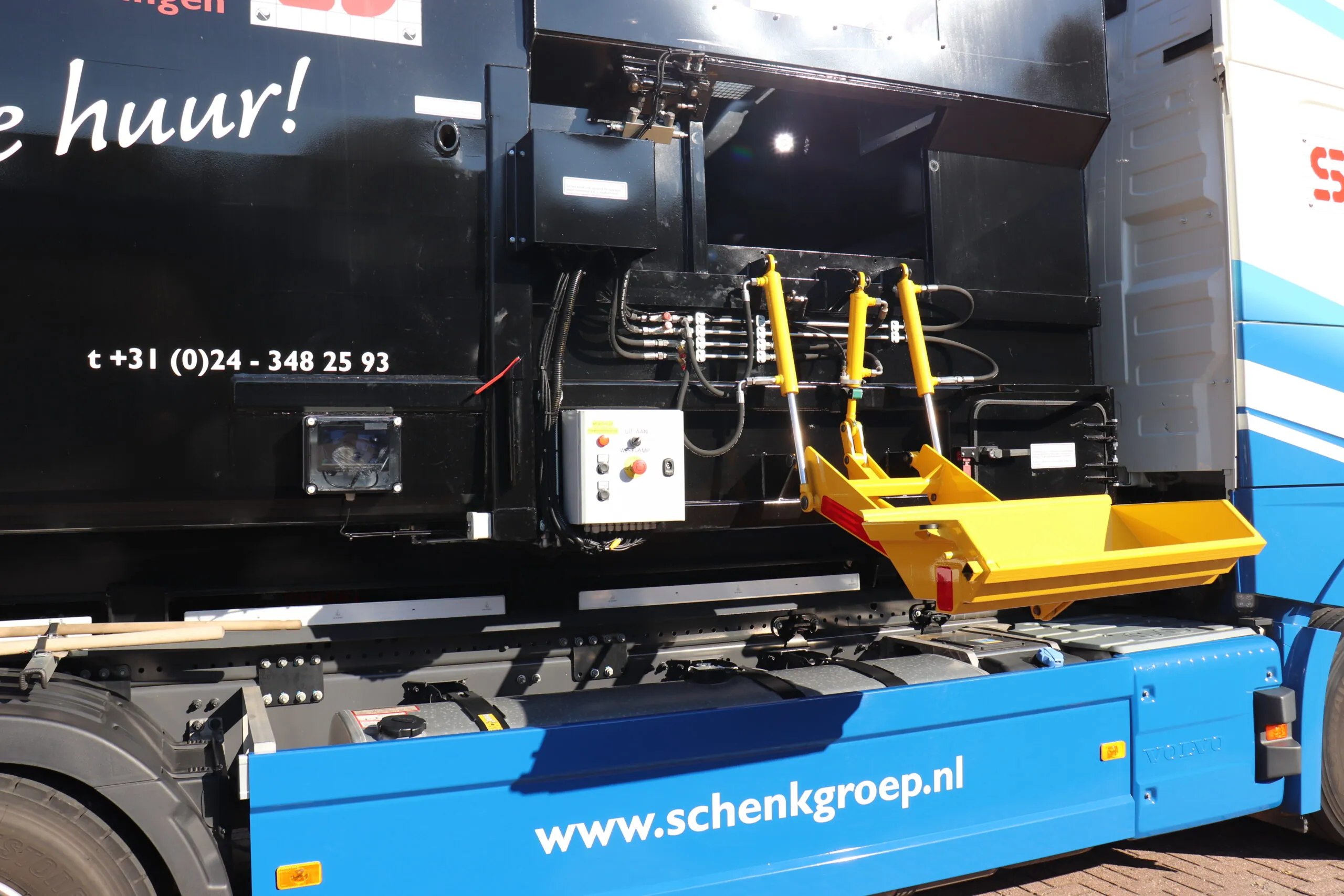 Gele en rode mechanische arm op een IPC-truck, in actie bij het beladen van afvalcontainers, met bedieningspaneel op de zijkant