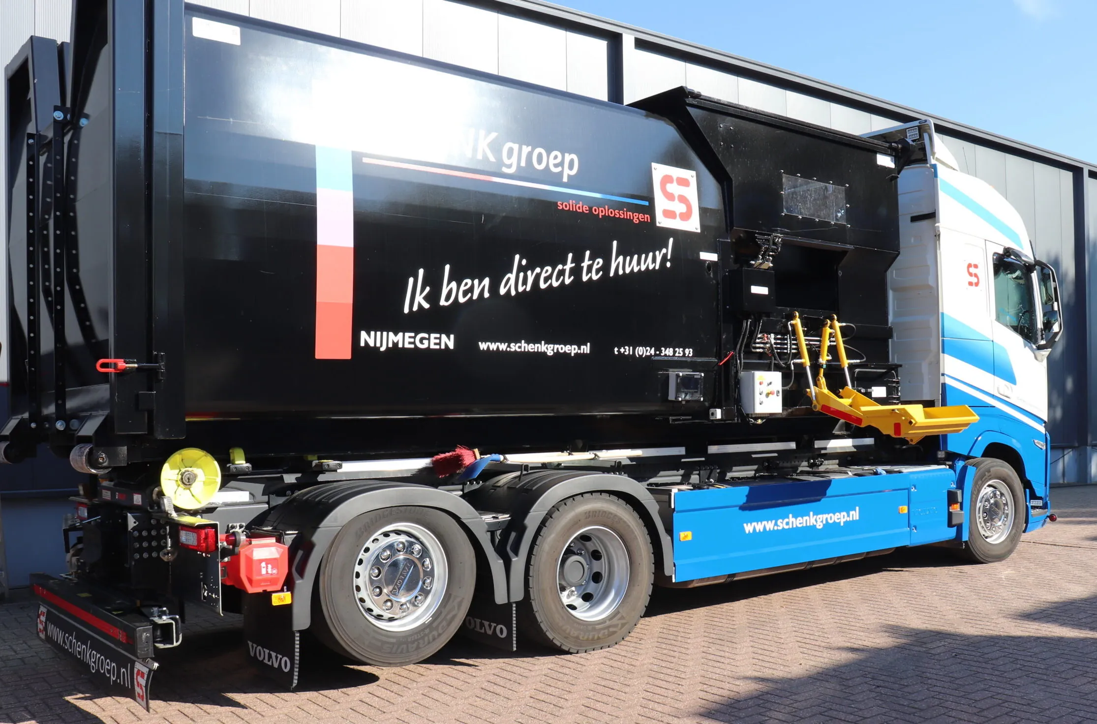 ommerciële truck met blauwe cabine en zwarte container, voorzien van Schenk Groep branding en verhuurtekst